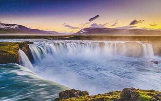 Der Godafoss bildet einen hufeisenförmigen Wasservorhang unter blauem Himmel.