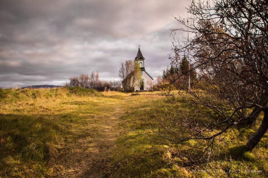 Thingvellir Church in Iceland with grassy path and autumn landscape under dramatic cloudy skies.