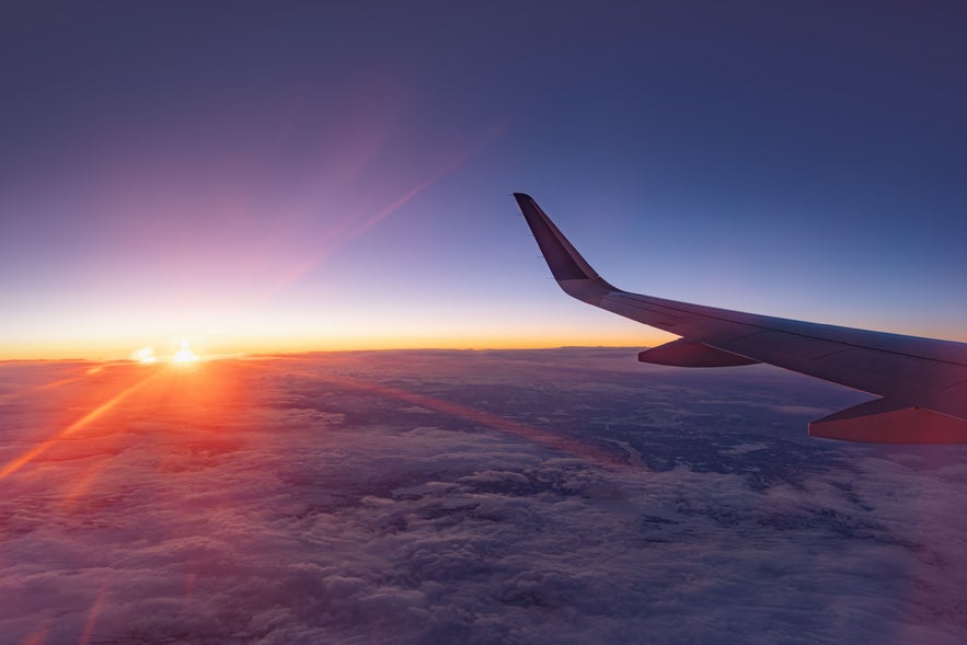 Airplane wing over clouds at sunrise, flying toward Iceland with snowy terrain below