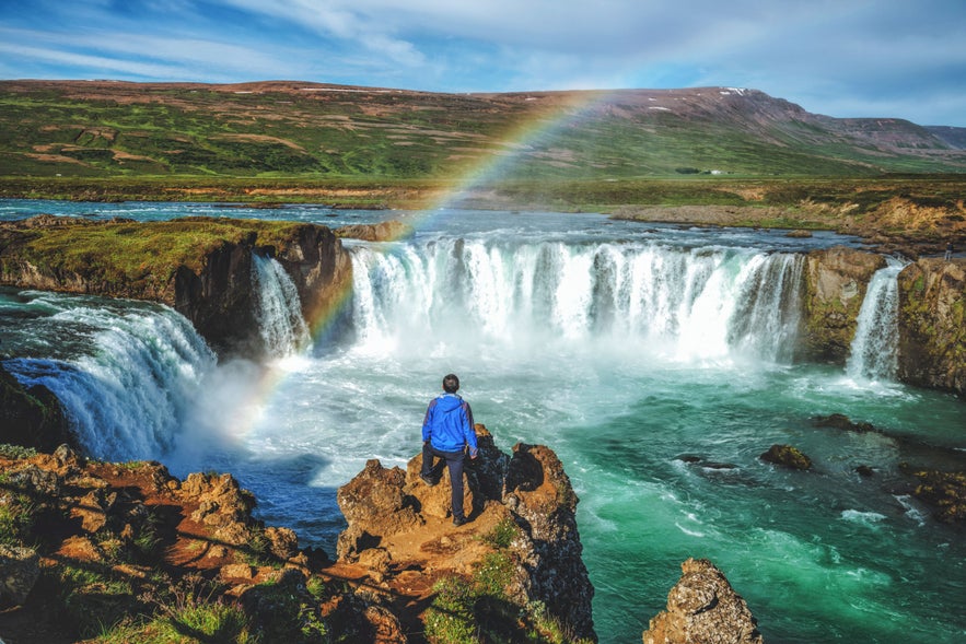Person soaking in the sun near Godafoss Waterfall in Iceland with a rainbow over the falls