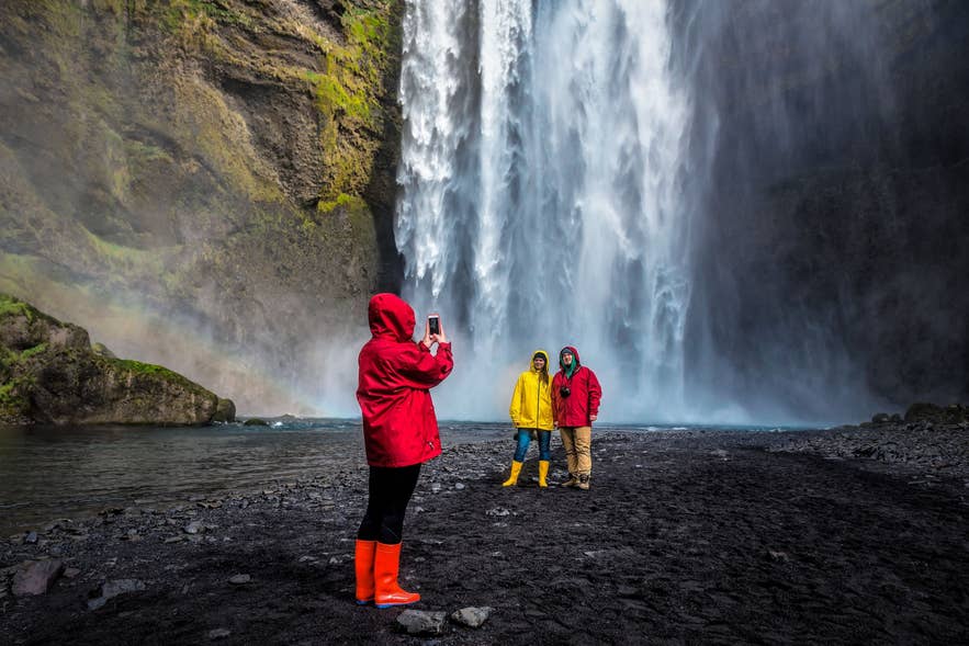 Travelers in rain jackets photograph Skogafoss Waterfall, on an Iceland South Coast trip that needs waterproofs every month.