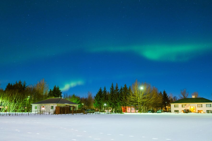 Northern lights over Hvolsvollur, South Iceland, with village homes and snowy fields along the South Coast at night.