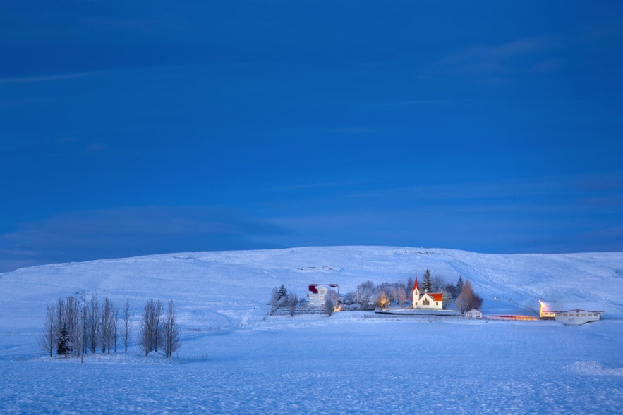 Snowy winter landscape near Hvolsvollur, South Iceland, with countryside homes, church, and rolling hills at dusk.