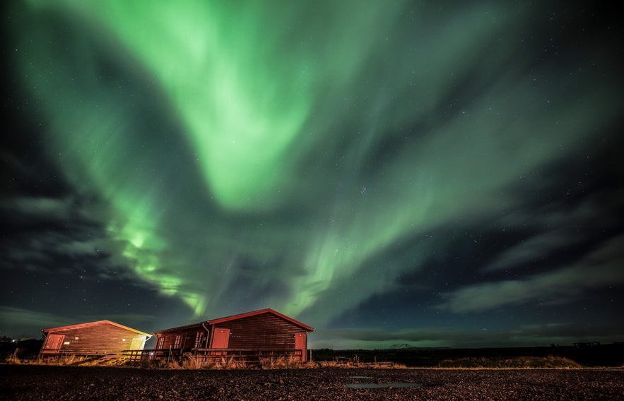 Northern lights dance over wooden cabins in Hella, Iceland, under a starry sky.