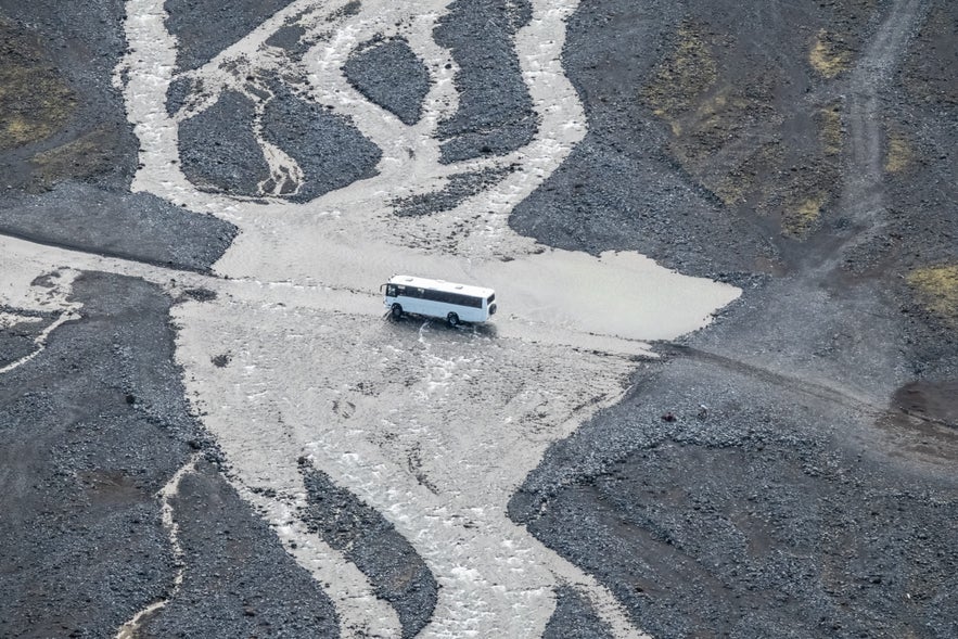 A white tour bus crosses a shallow glacial river on a rocky highland road in Iceland.