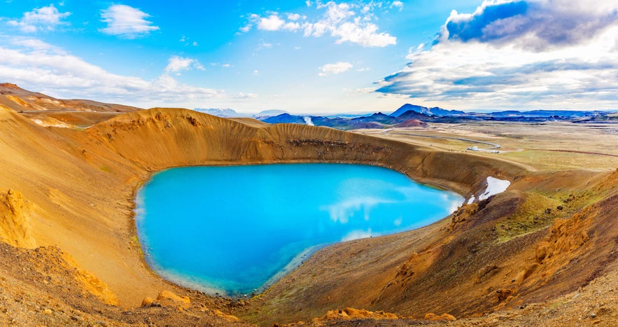 Bright blue lake fills Viti Crater in Krafla, surrounded by orange-brown volcanic terrain under a partly cloudy sky.