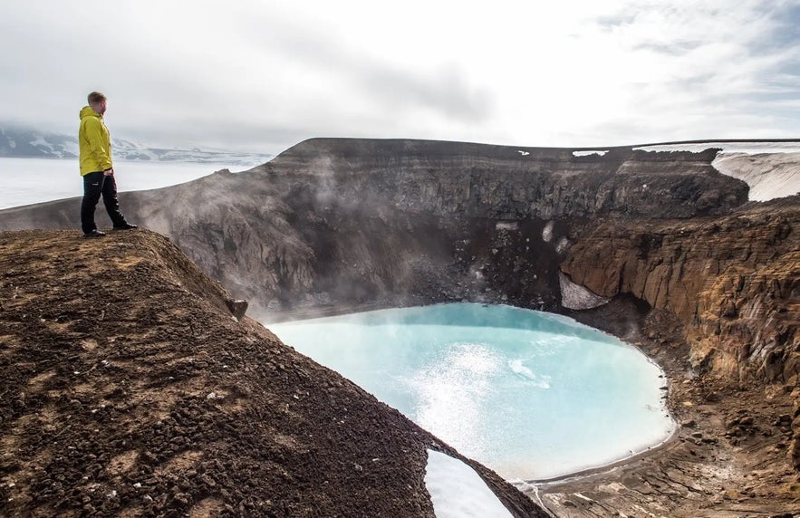 A man in a yellow jacket stands on the edge of Askja Caldera, gazing over the milky-blue Viti Crater Lake in Iceland's Highlands.