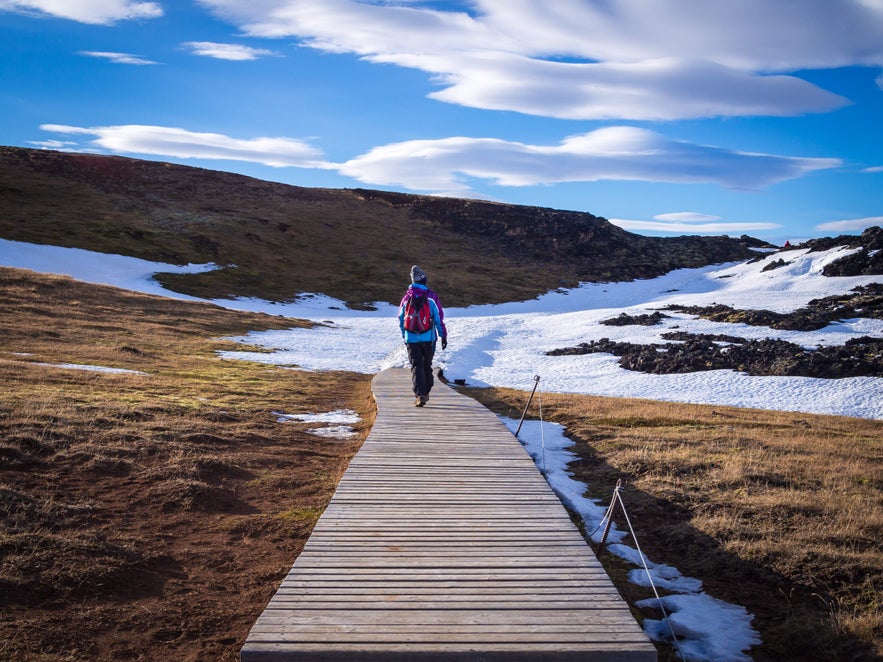 A hiker walks along a wooden boardwalk through melting snow and lava fields in the Krafla Volcanic Area.