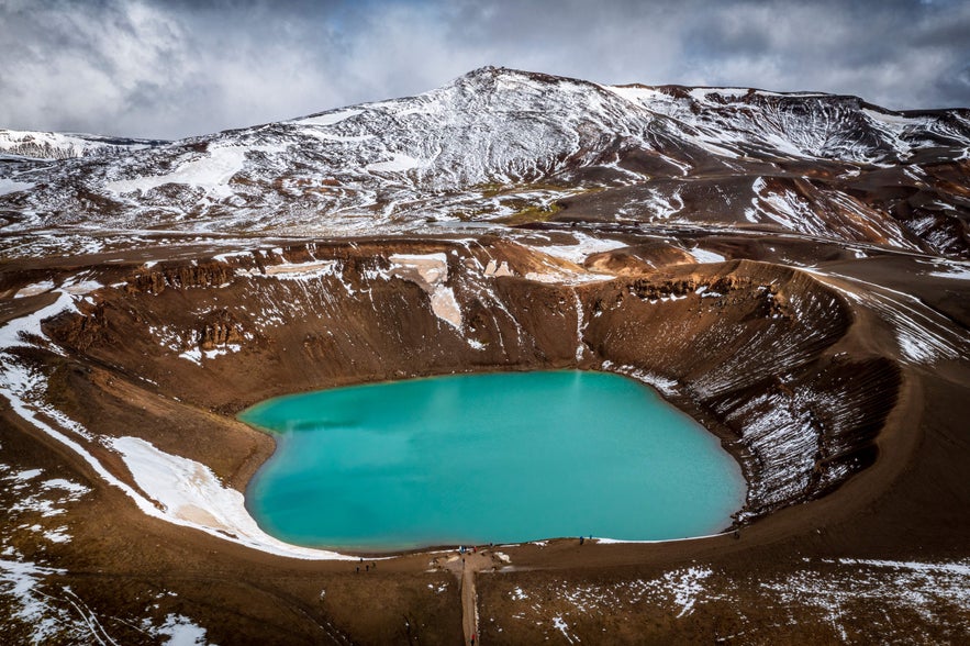 Aerial view of Viti Crater in Krafla, Iceland, with turquoise water surrounded by snow-dusted volcanic slopes and dark clouds.