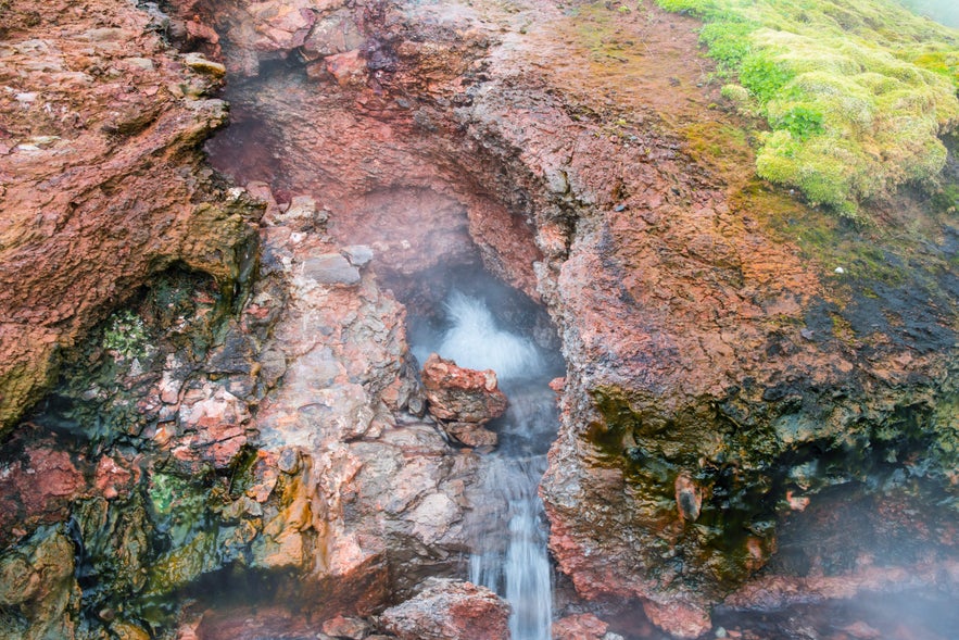 A geothermal spring bursts from red volcanic rock at Deildartunguhver, surrounded by moss and minerals.