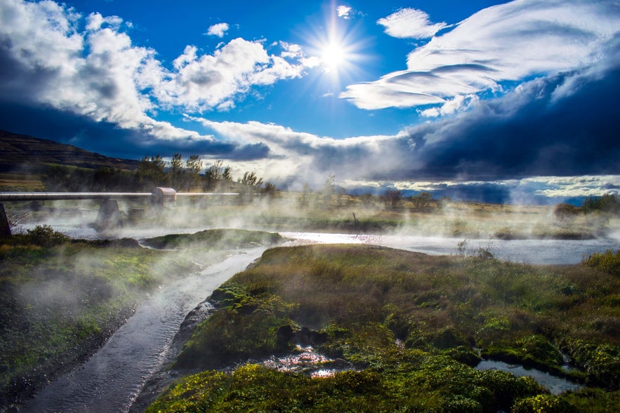Hot water streams through grassy fields at Deildartunguhver as sun rays break through clouds and mist.
