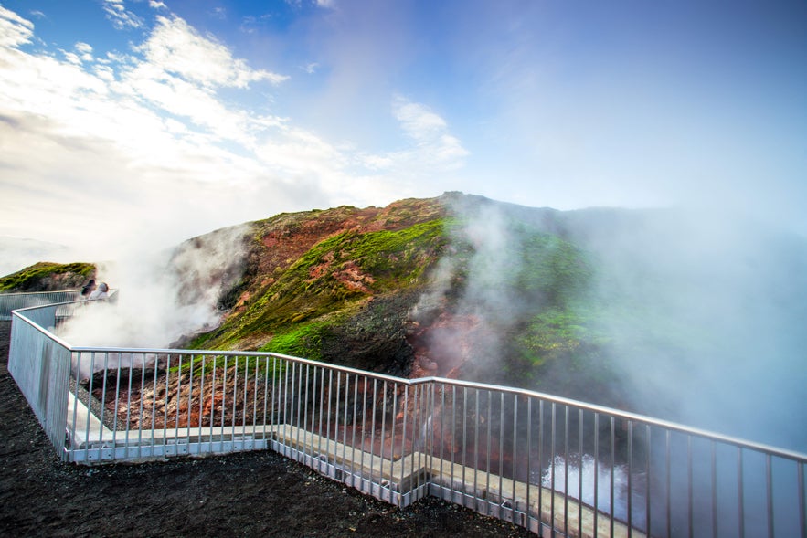 Steam rises from Deildartunguhver&rsquo;s fenced geothermal area, with mossy red slopes and observation platforms.