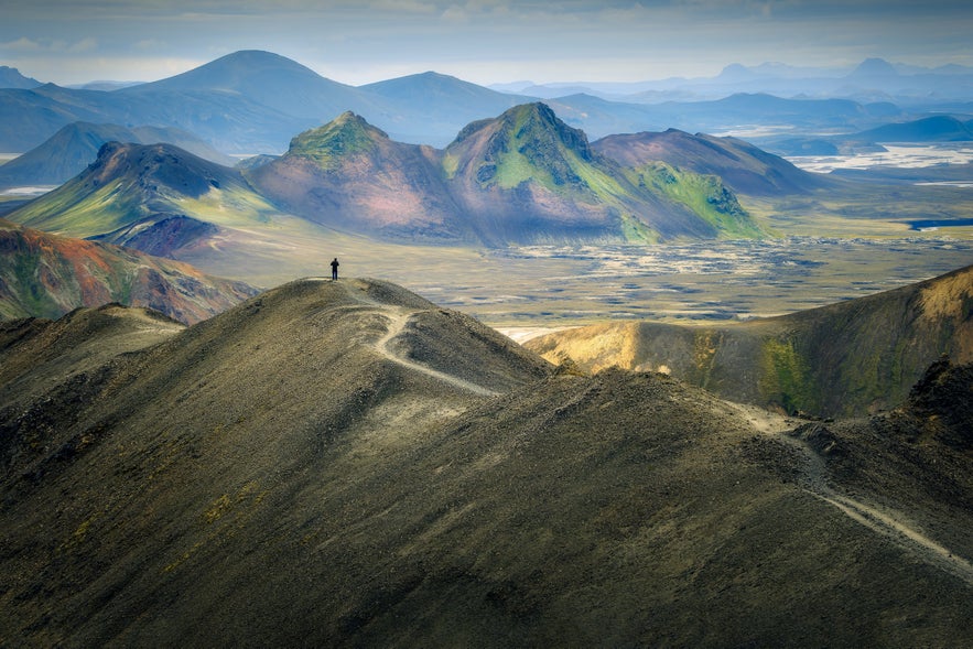 A hiker standing on a ridge with panoramic views of Landmannalaugar&rsquo;s colorful mountains.