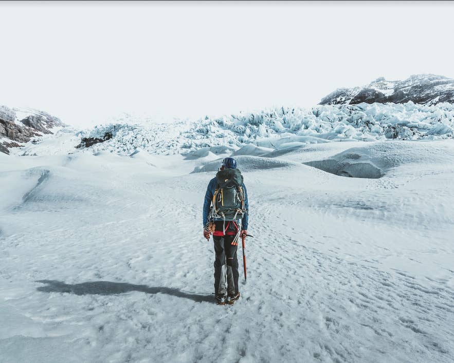 A hiker stands on a glacier in Iceland, facing blue ice ridges while holding an ice axe and wearing a backpack. A hiker stands on a glacier in Iceland, facing blue ice ridges while holding an ice axe and wearing a backpack.