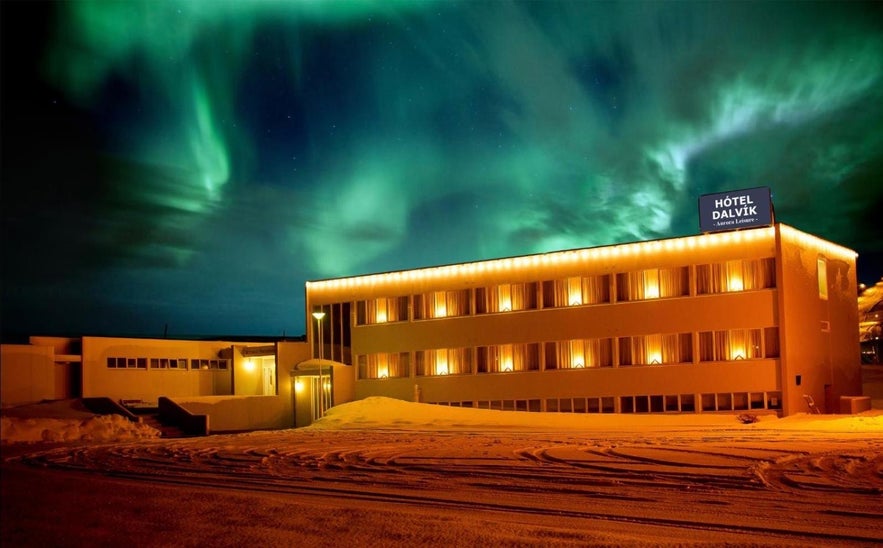 Hotel Dalvik in North Iceland lit up at night with the northern lights glowing above and fresh snow covering the ground.
