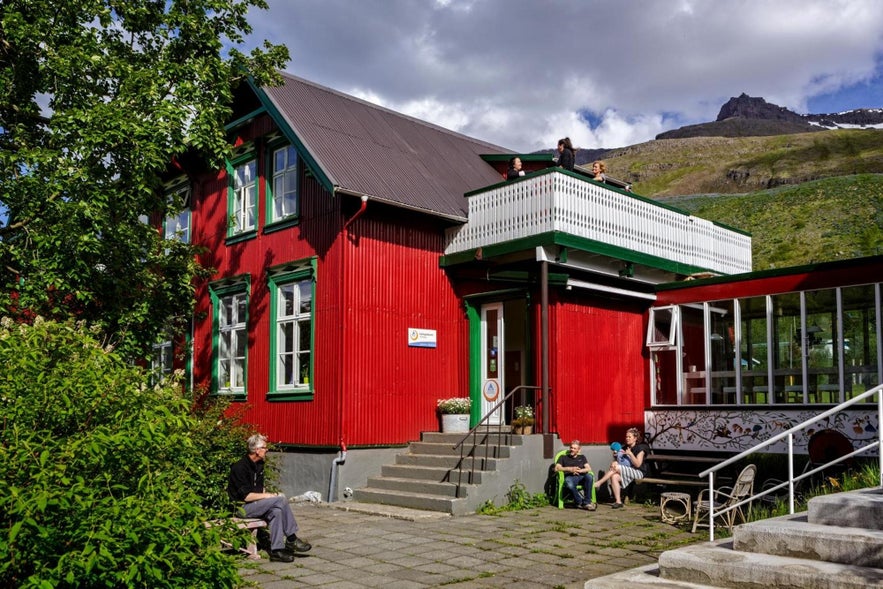 A red and green wooden building housing Hafaldan HI Hostel sits beneath a mountain in East Iceland. Guests relax outside in the sun.