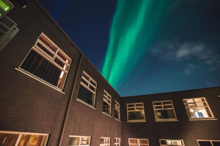 Northern lights dance above the courtyard of a dark-colored hotel building, with glowing windows illuminating the night in Iceland.