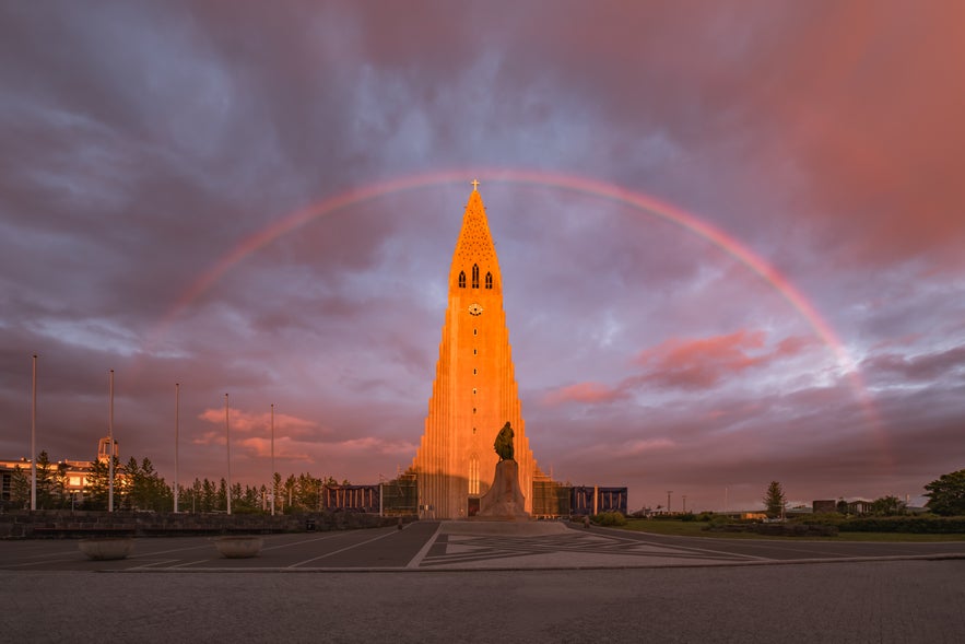 The Hallgr&iacute;mskirkja church in Reykjav&iacute;k glows at sunset with a rainbow arching over it under a dramatic pink and purple sky.