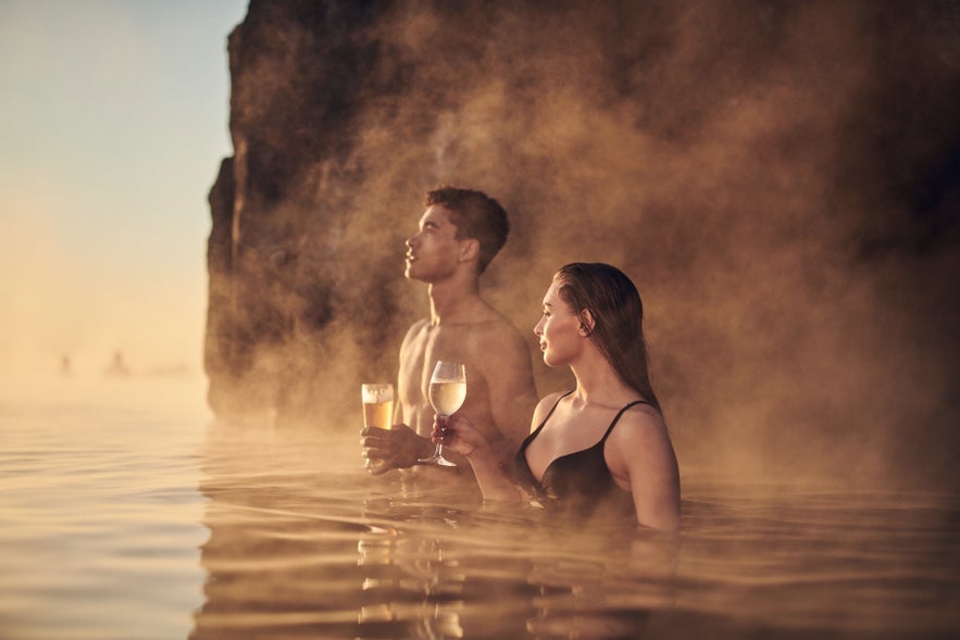 A couple relaxing in Sky Lagoon at sunset, enjoying drinks as steam rises from the warm geothermal water.