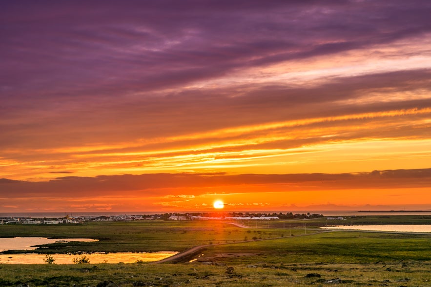 The midnight sun is setting over Reykjavik, with warm orange light reflecting on ponds and fields beneath a purple sky.