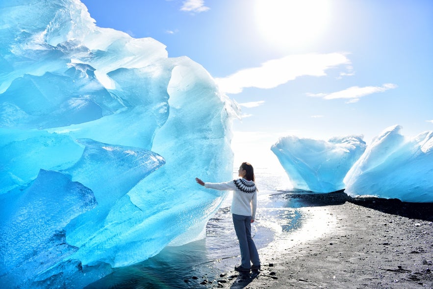Traveler in Iceland standing beside a large blue iceberg on the black sand Diamond Beach.