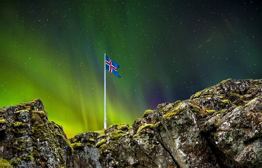 The northern lights glow above the Icelandic flag as it stands over the believed location of Logberg Law Rock, surrounded by rocks.