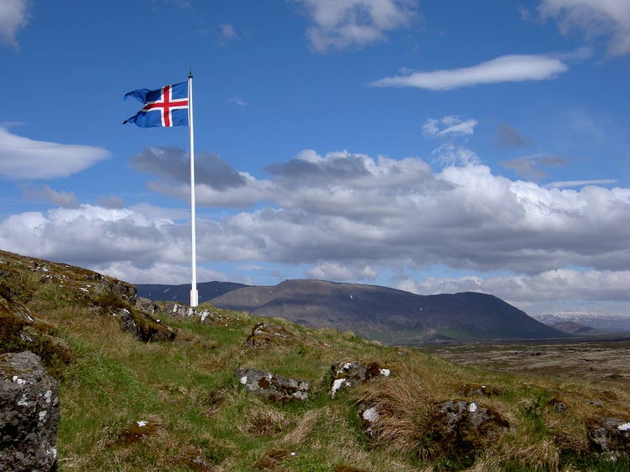 The Icelandic flag waves above green grass and scattered rocks with mountains rising in the distance near Logberg Law Rock.