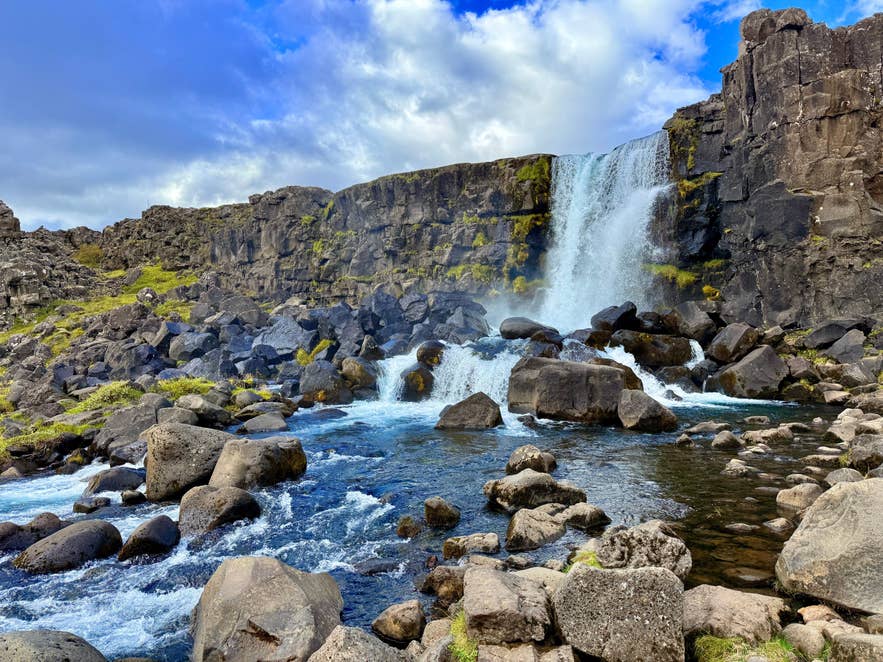 Oxararfoss Waterfall cascades over mossy cliffs beneath a bright blue sky.