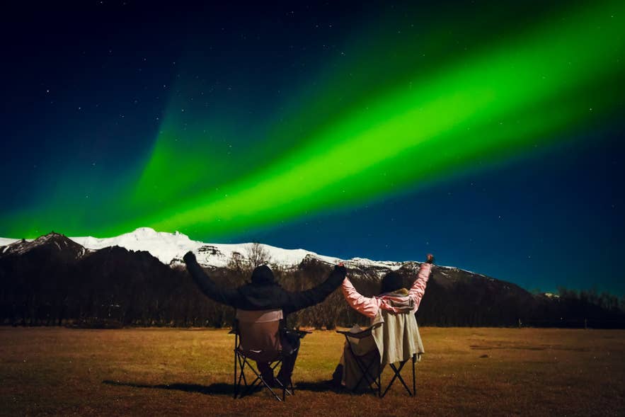 Par ser på nordlyset på Island, sittende ute under grønn aurora og snødekte fjell om natten.