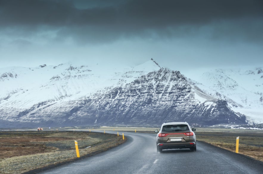 Car driving along a scenic road in Iceland, with snow-covered mountains rising under dark clouds in the background.