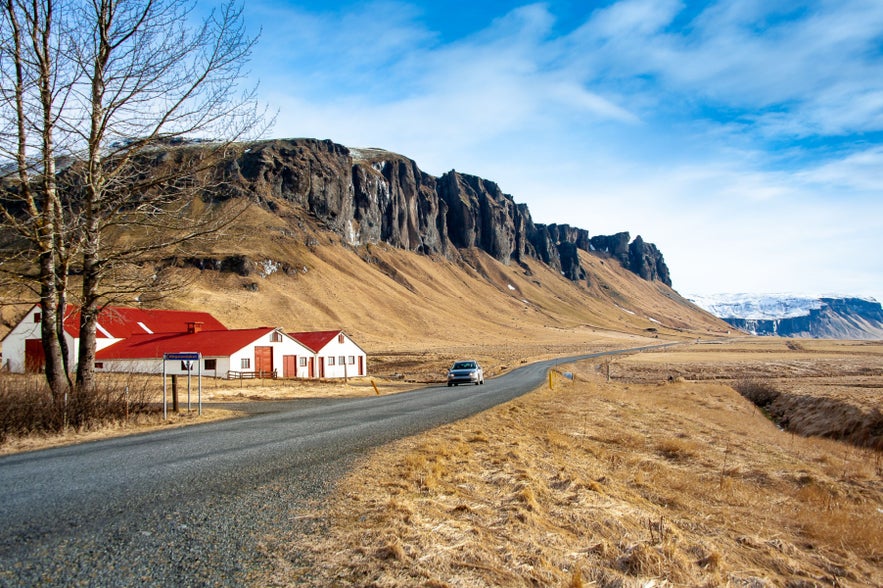Iceland&rsquo;s Ring Road winding past red-roofed houses and dramatic cliffs, showcasing a scenic drive through rural Iceland.