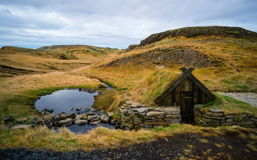 Hrunalaug natuurlijke warmwaterbron bij Fludir, Zuid-IJsland, met een stenen bad en een turfhuisje in een groene vallei.