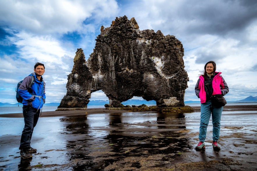 Travelers standing near Hvitserkur Sea Stack, a unique basalt rock formation resembling a drinking dragon in North Iceland.