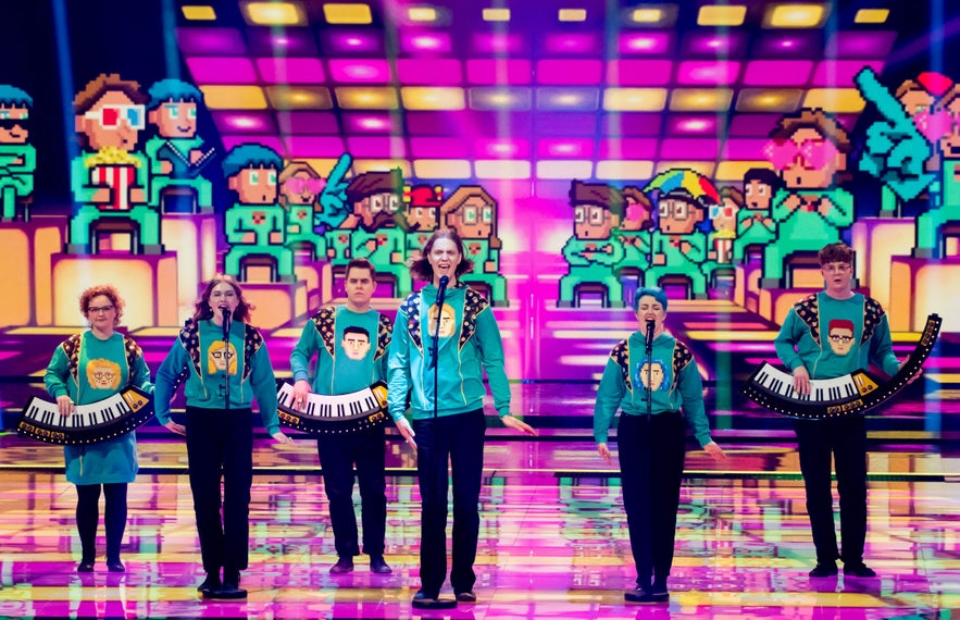 Da&eth;i og Gagnamagni&eth; performing "10 Years" at Eurovision for Iceland, wearing signature teal sweaters with pixel-art portraits and holding custom curved keyboards.