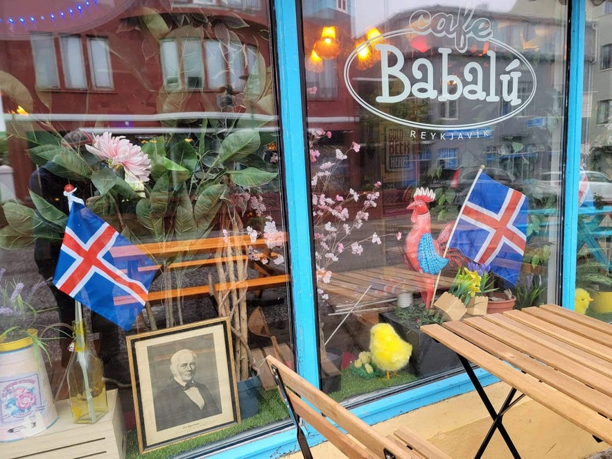 Colorful window display of Cafe Babalu in Reykjavik featuring Icelandic flags, rooster figurine, flowers, and eclectic decorations.