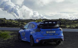A blue rally car parked beside steaming geothermal vents in Hvalfjordur near Reykjavik.