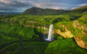 Luftaufnahme des Seljalandsfoss-Wasserfalls, der von einer Klippe in ein grünes Tal mit Wanderwegen darunter stürzt.