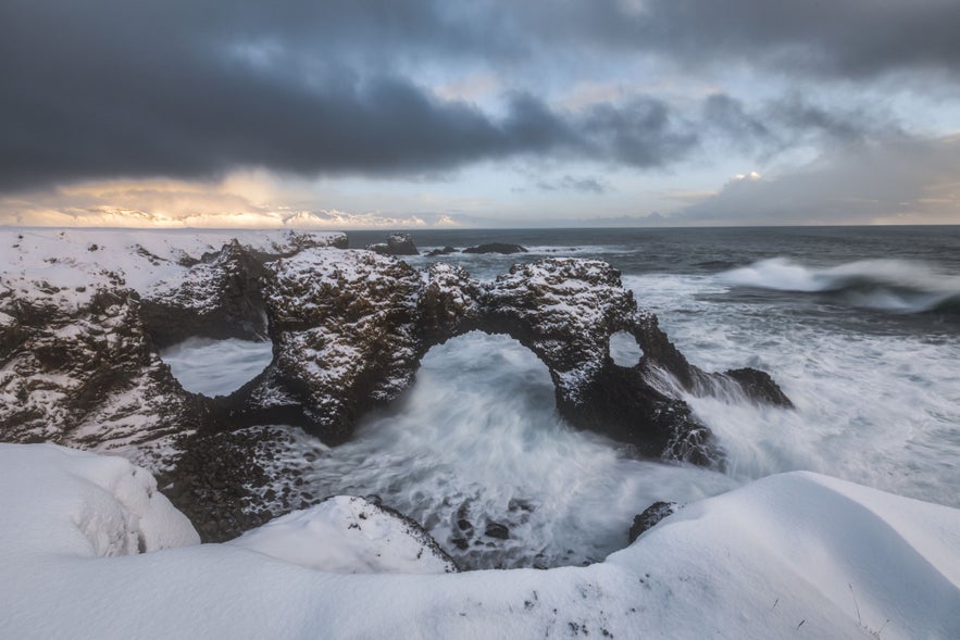 Snow-covered Gatklettur Rock Arch in Arnarstapi, Snaefellsnes Peninsula, Iceland, with Atlantic waves crashing in winter.