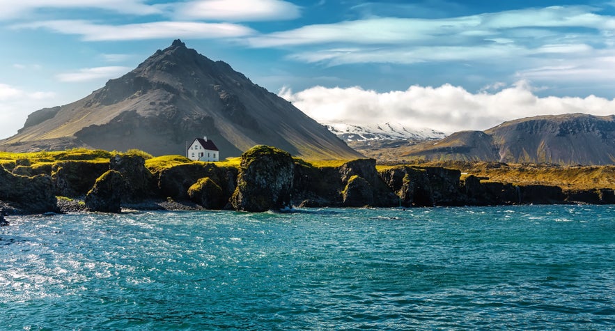 White house on the rocky coast of Arnarstapi with a mountain backdrop and blue ocean under a bright sky on the Sn&aelig;fellsnes Peninsula.