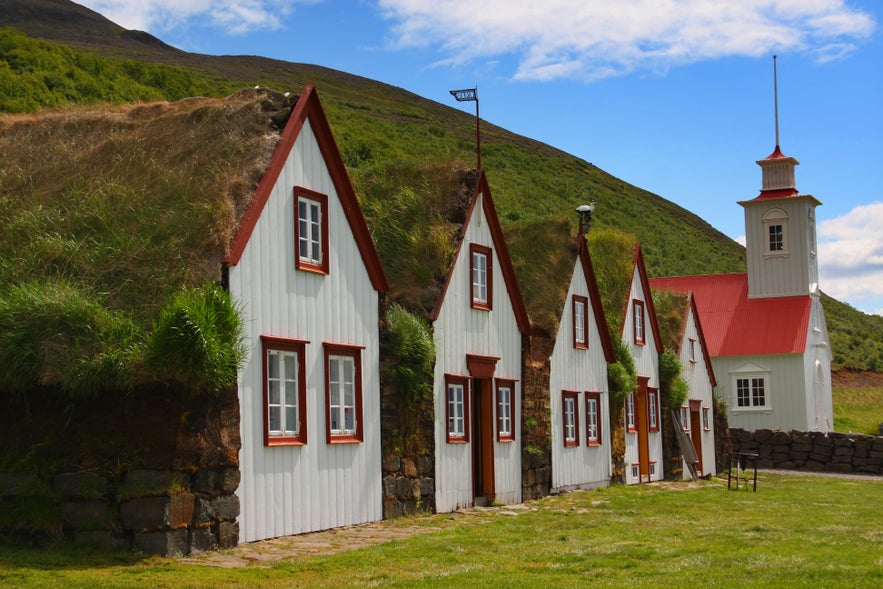 Traditional Icelandic turf houses at Laufas farm with white wooden gables, grass-covered roofs, and a red-roofed country church in the background. Traditional Icelandic turf houses at Laufas farm with white wooden gables, grass-covered roofs, and a red-roofed country church in the background.