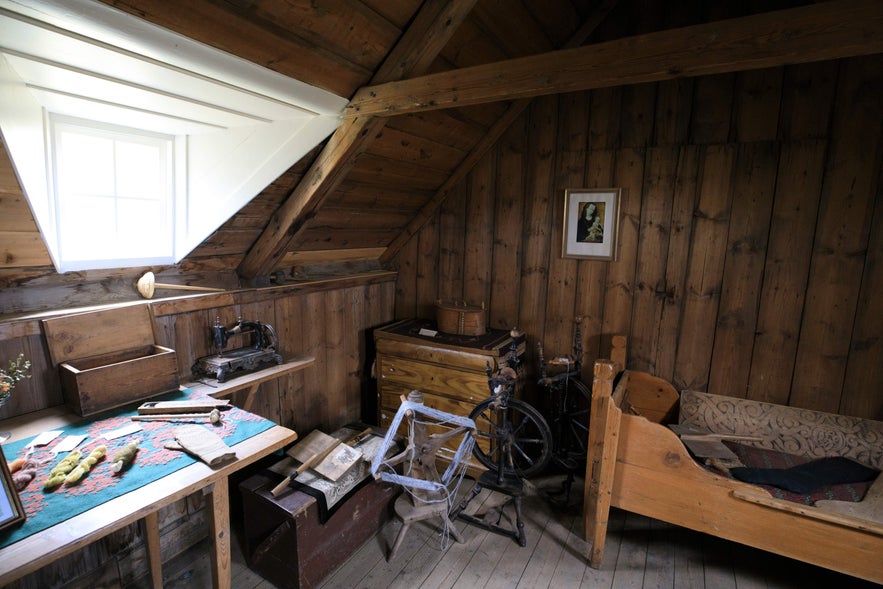 Interior of a traditional Icelandic turf house at Laufas Folk Museum, featuring wood-paneled walls, a small attic window, and historic artifacts like a spinning wheel and antique bed.