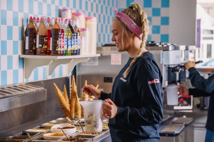 Staff preparing a mix-in ice cream (brag&eth;arefur) at &Iacute;sb&uacute;&eth; Huppu in Reykjavik with cones and toppings on the counter
