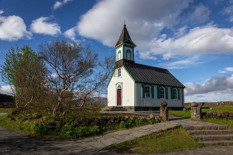 A pathway and steps lead to Thingvellir Church under a bright, cloudy summer sky.