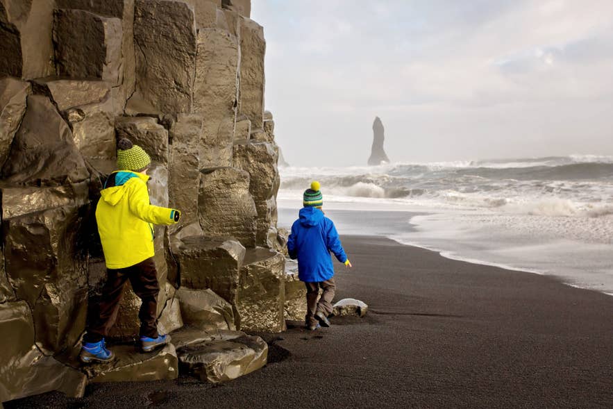IJsland met kinderen die het zwarte zandstrand Reynisfjara bij Vik verkennen, met de Reynisdrangar zeestapels op de achtergrond.