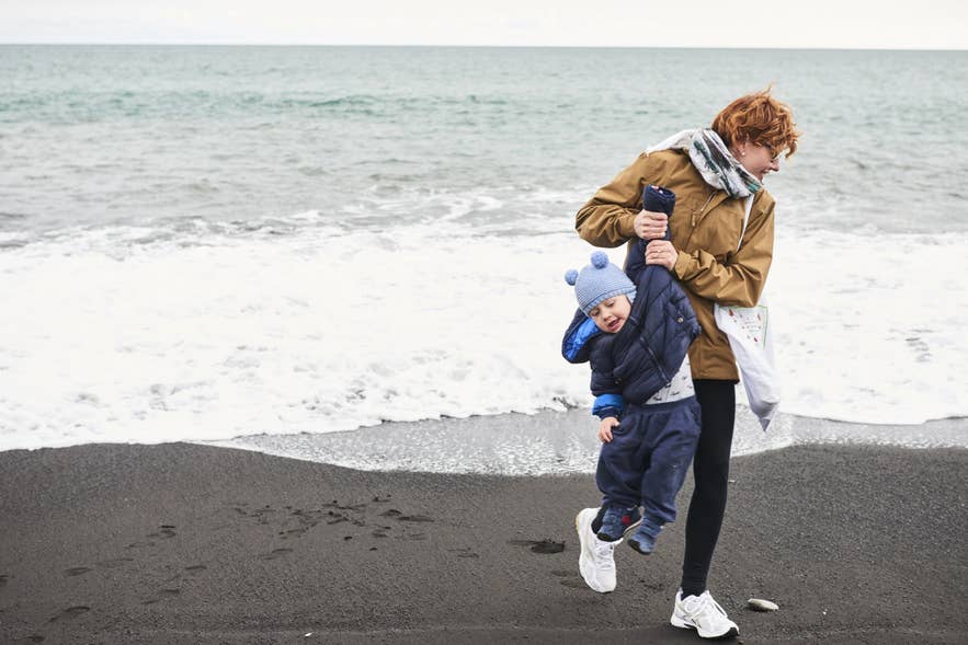 Ouder draagt kind op een zwart zandstrand bij Vík, genietend van IJsland met kinderen langs de zuidkust.