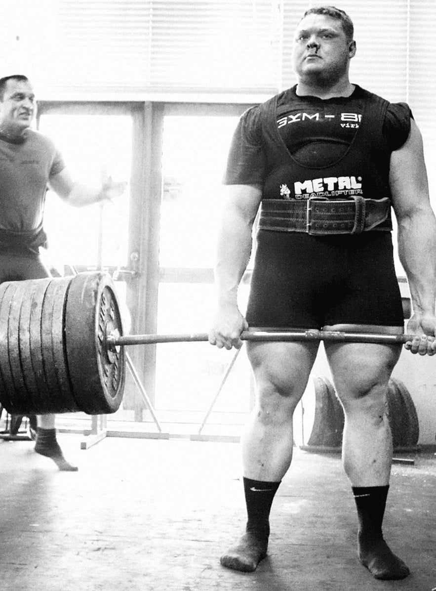 Benedikt Magnusson performs a heavy deadlift in a gym, gripping a heavily loaded barbell while a coach watches in the background.