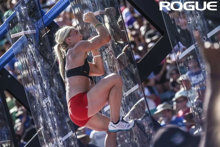 Katrin Davidsdottir climbs a pegboard during a CrossFit Games competition, gripping wooden pegs as a crowd watches in the background.