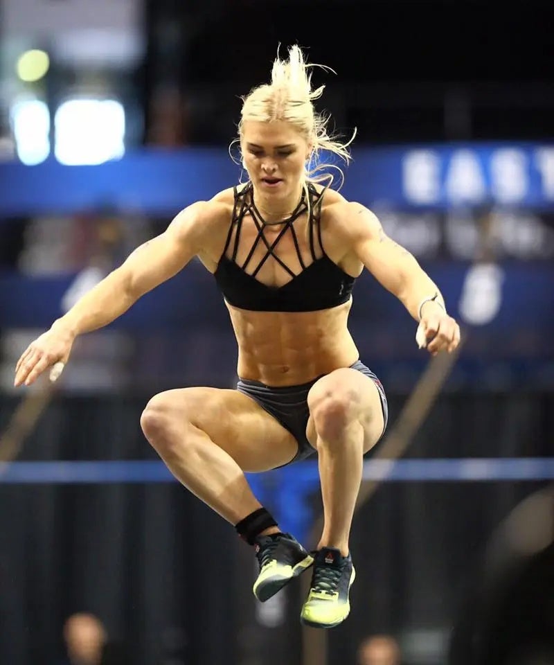 Katrin Tanja Davidsdottir performs a jump during a CrossFit competition, mid-air with knees bent and arms extended for balance inside an arena.