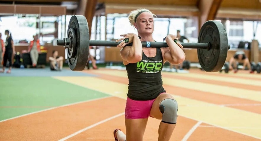 Ragnheidur Sara Sigmundsdottir performs a barbell front rack lunge during a CrossFit training session inside an indoor sports hall.