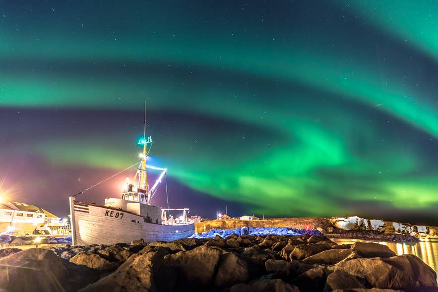 Green northern lights glowing above a fishing boat in an Icelandic harbor, with rocks and village lights along the shoreline at night.